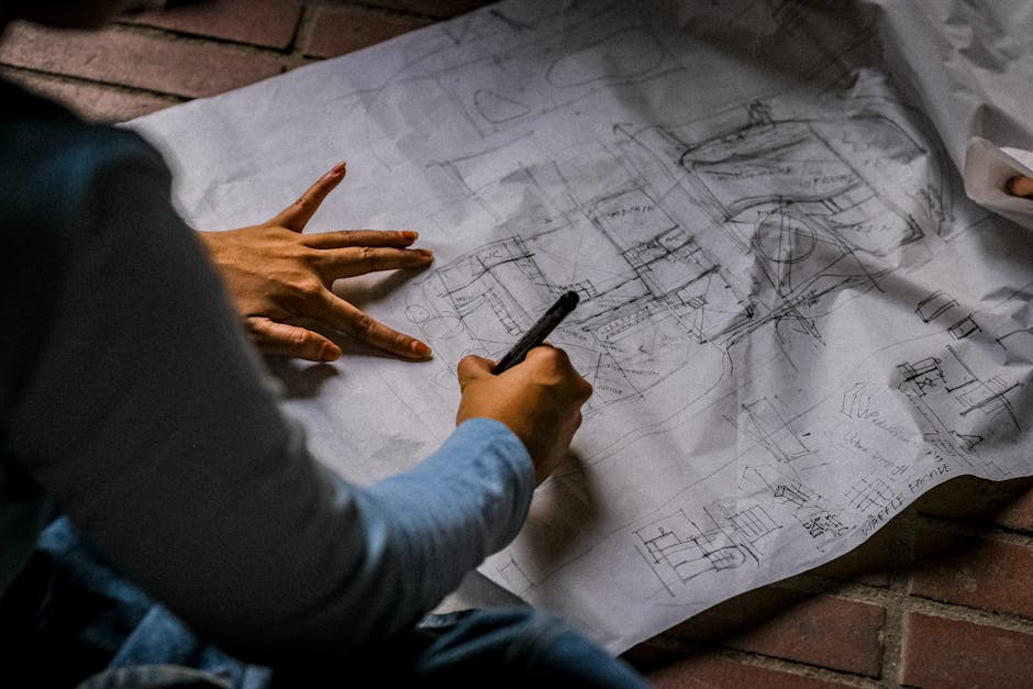 Person sketching architectural design on blueprint paper with pen on brick floor.
