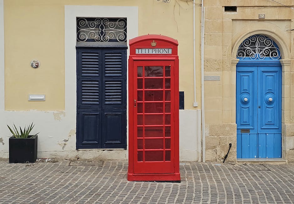 Classic red telephone booth on a charming Maltese street with colorful doors.